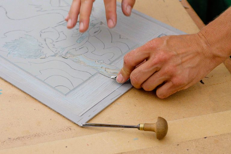 Two hands carving away lino with tools to create a pattern to print with.