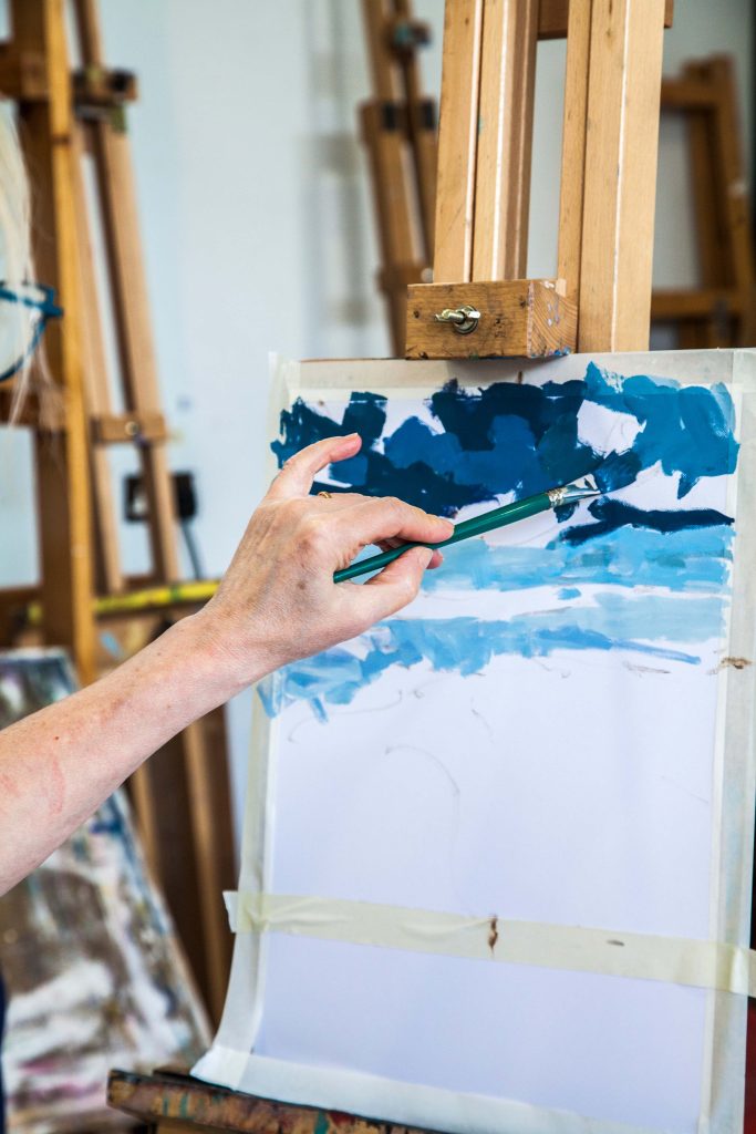 A hand painting the sky scene of a painting on an easels.
