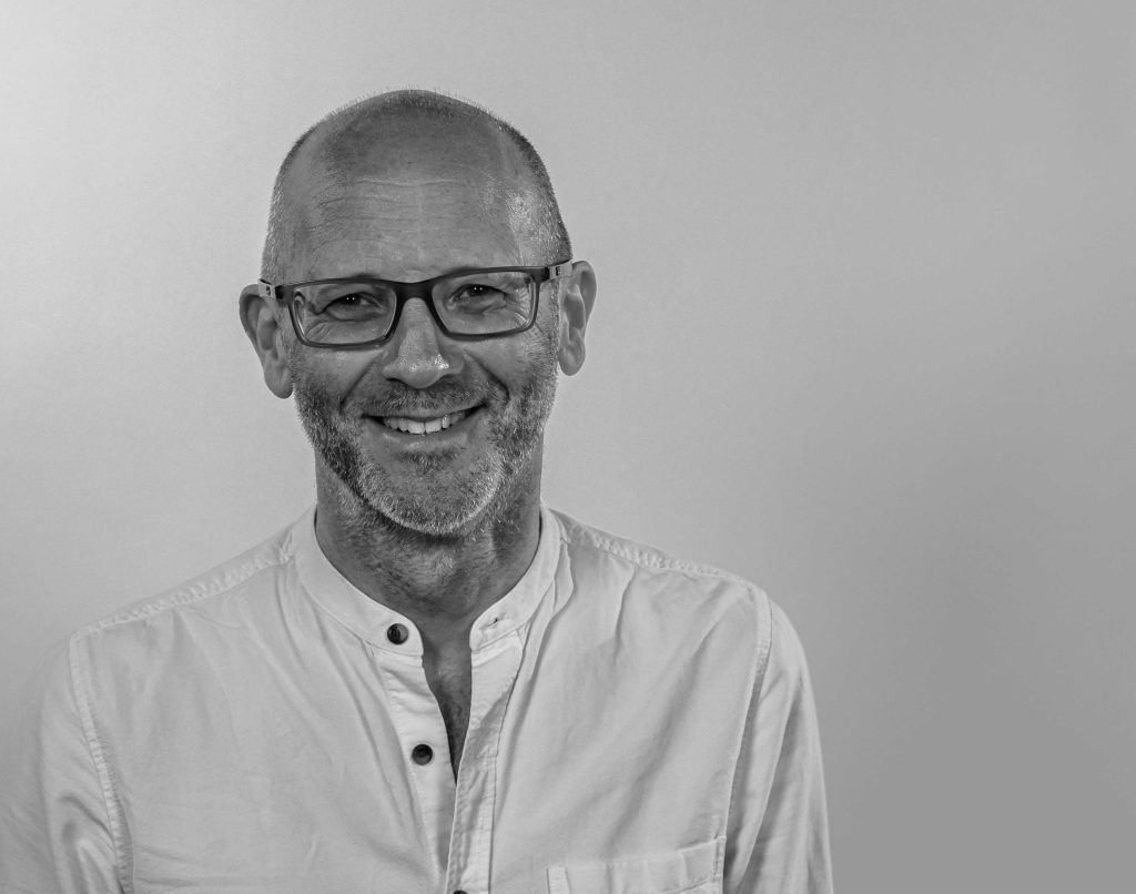 A monochromatic photo of a bald white man with glasses, smiling.