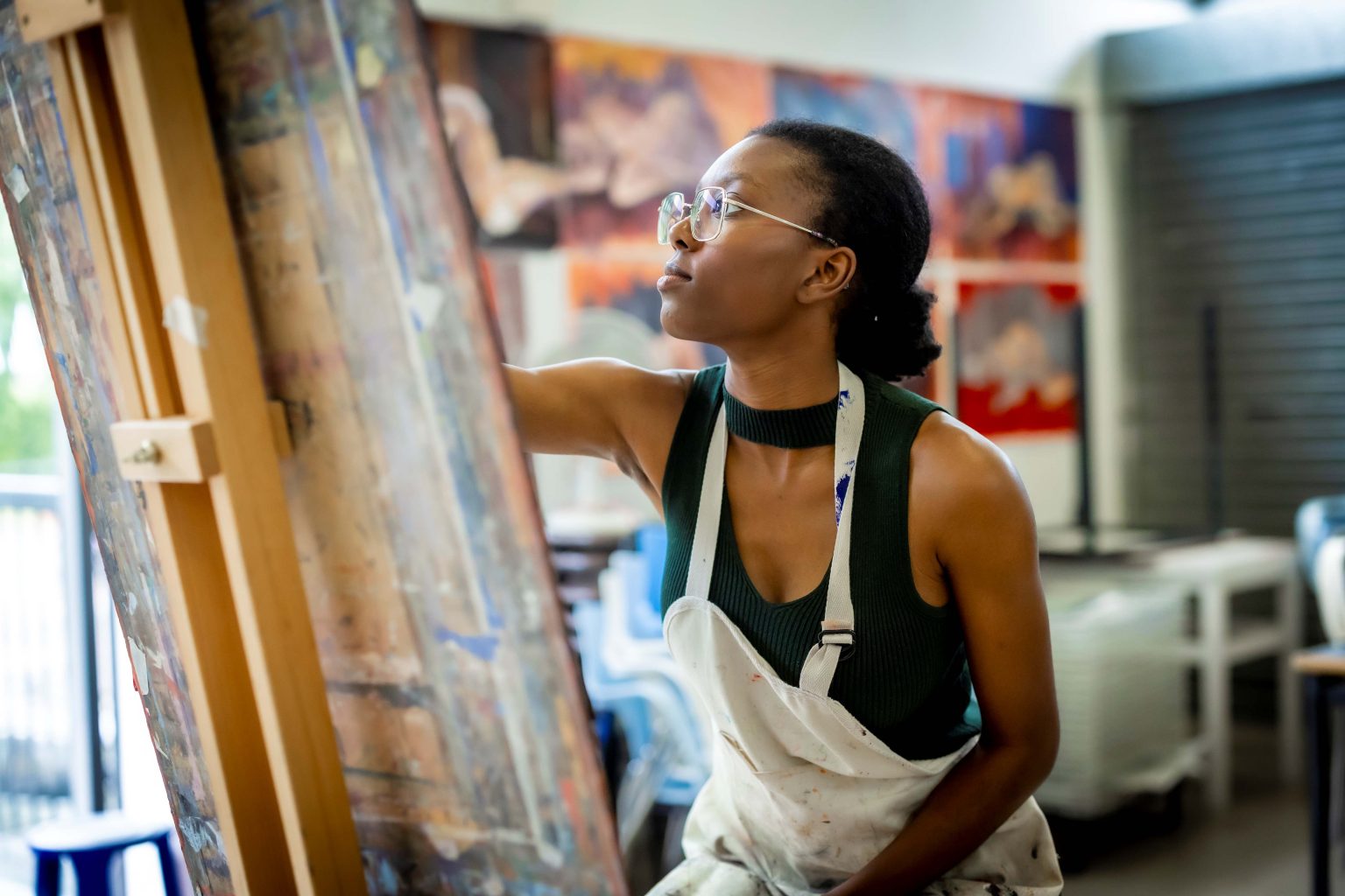 A Black woman painting on an easel while wearing an apron. Behind her are paintings of life models.