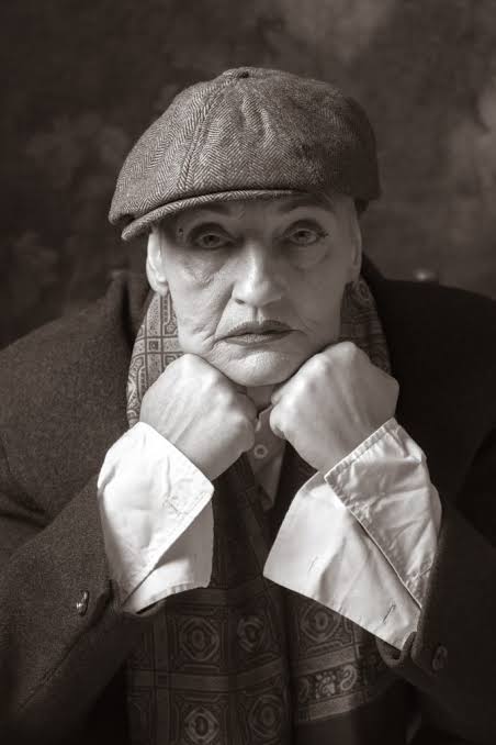 An older white woman with a shaved head and red lipstick, resting her chin on her hand while wearing a flat cap and suit.
