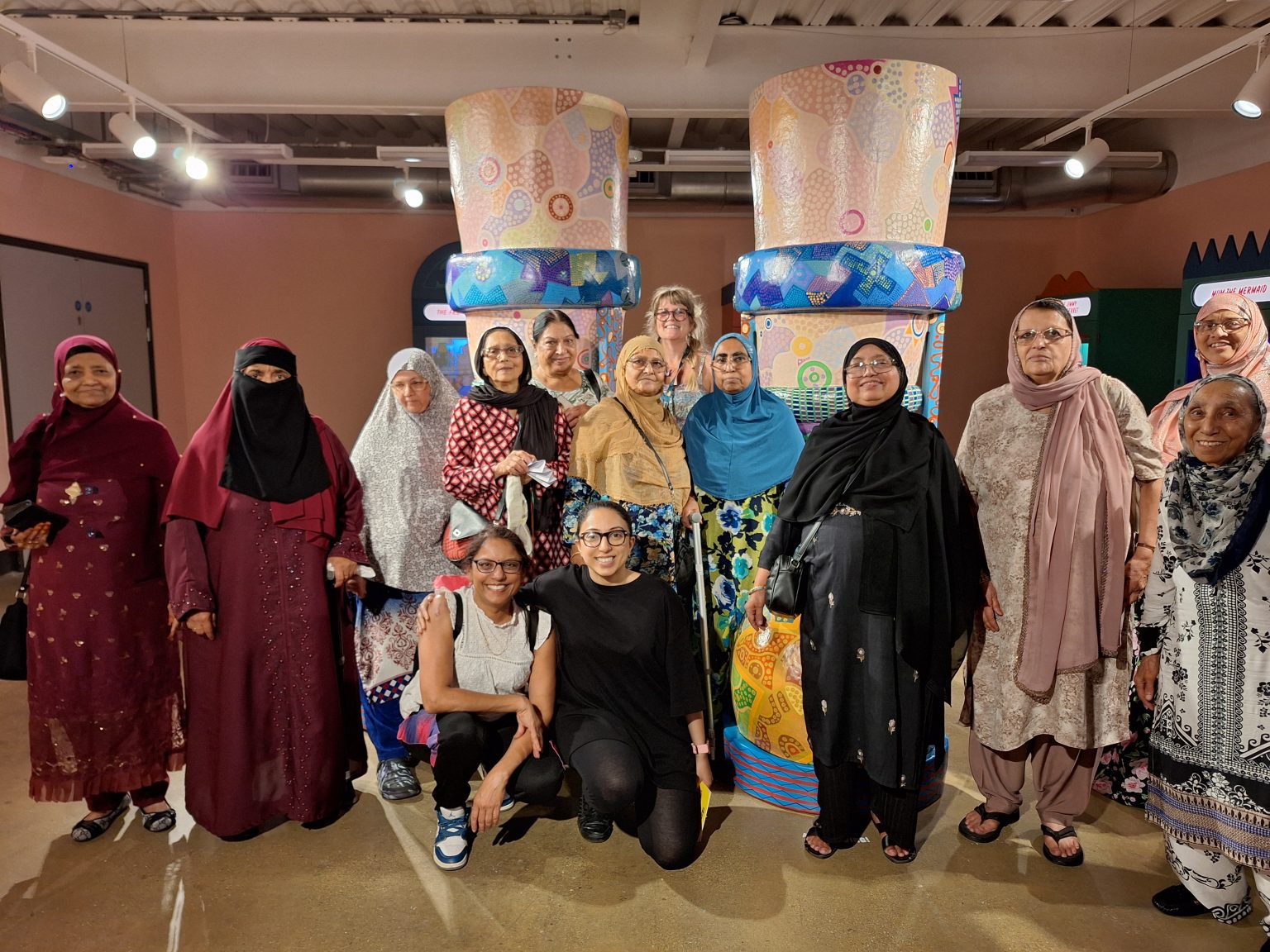 A group of women stand together in a gallery in front of a huge pair of painted legs and shoes.