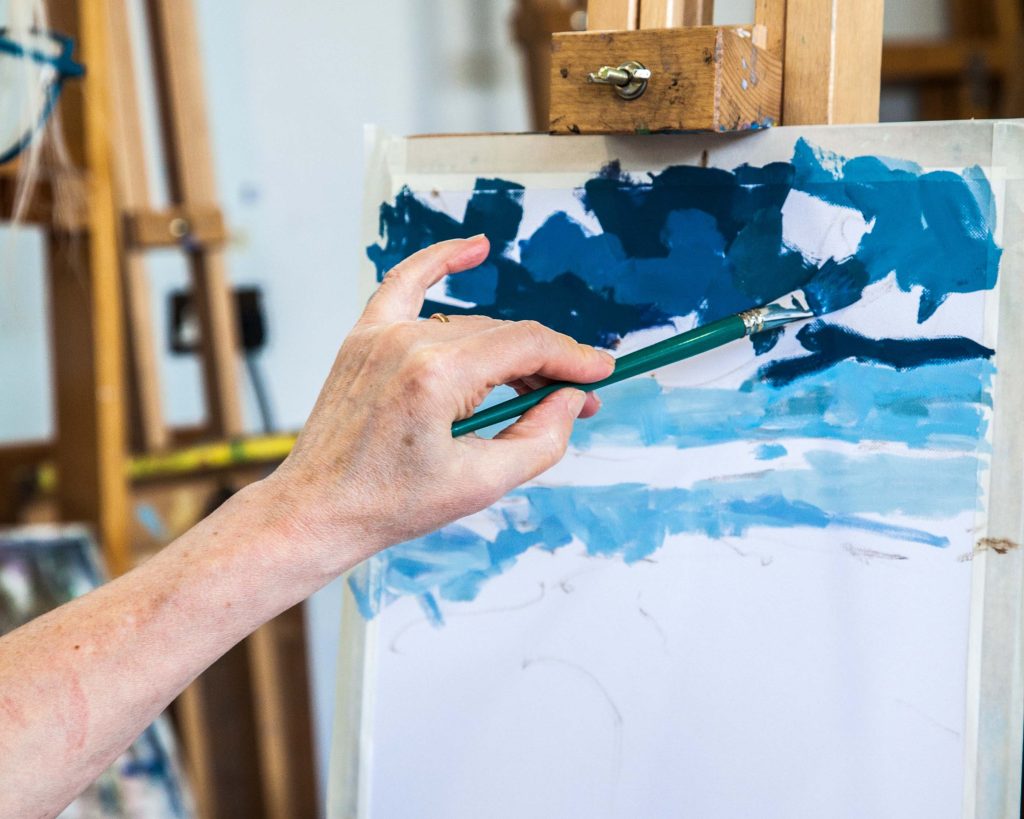 A hand painting the sky scene of a painting on an easels.