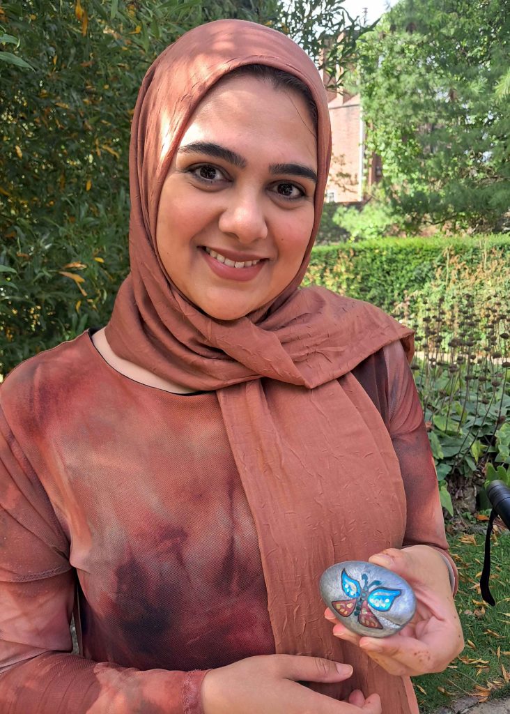 A woman wearing a hijab smiling and holding a pebble she has drawn a butterfly onto.
