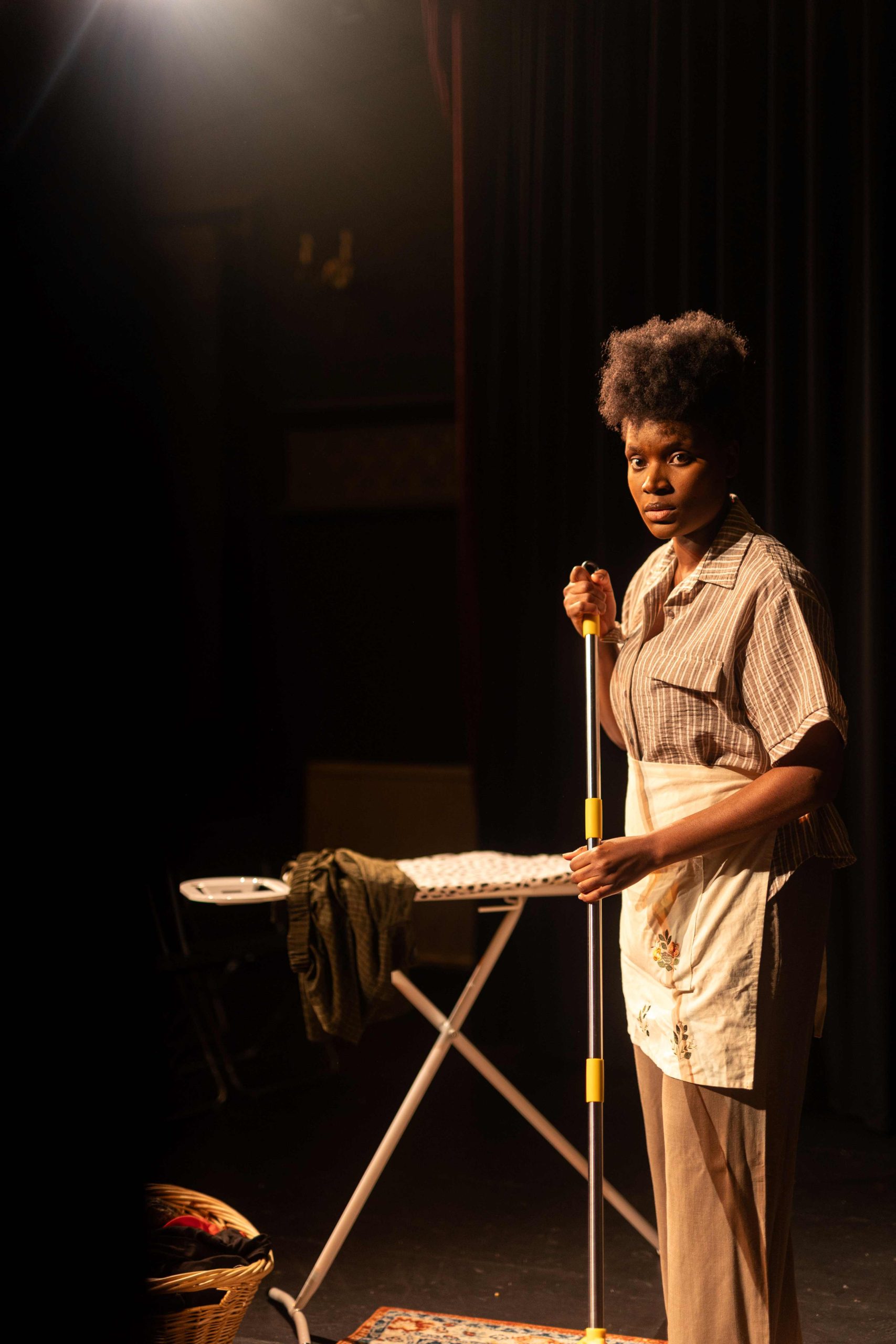 A black woman stood holding a mop in front of a ironing board.