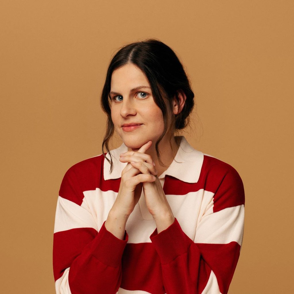 A white woman with brown hair in an up-do, wearing a striped red and white polo top. She has her hands underneath her chin with a brown background.