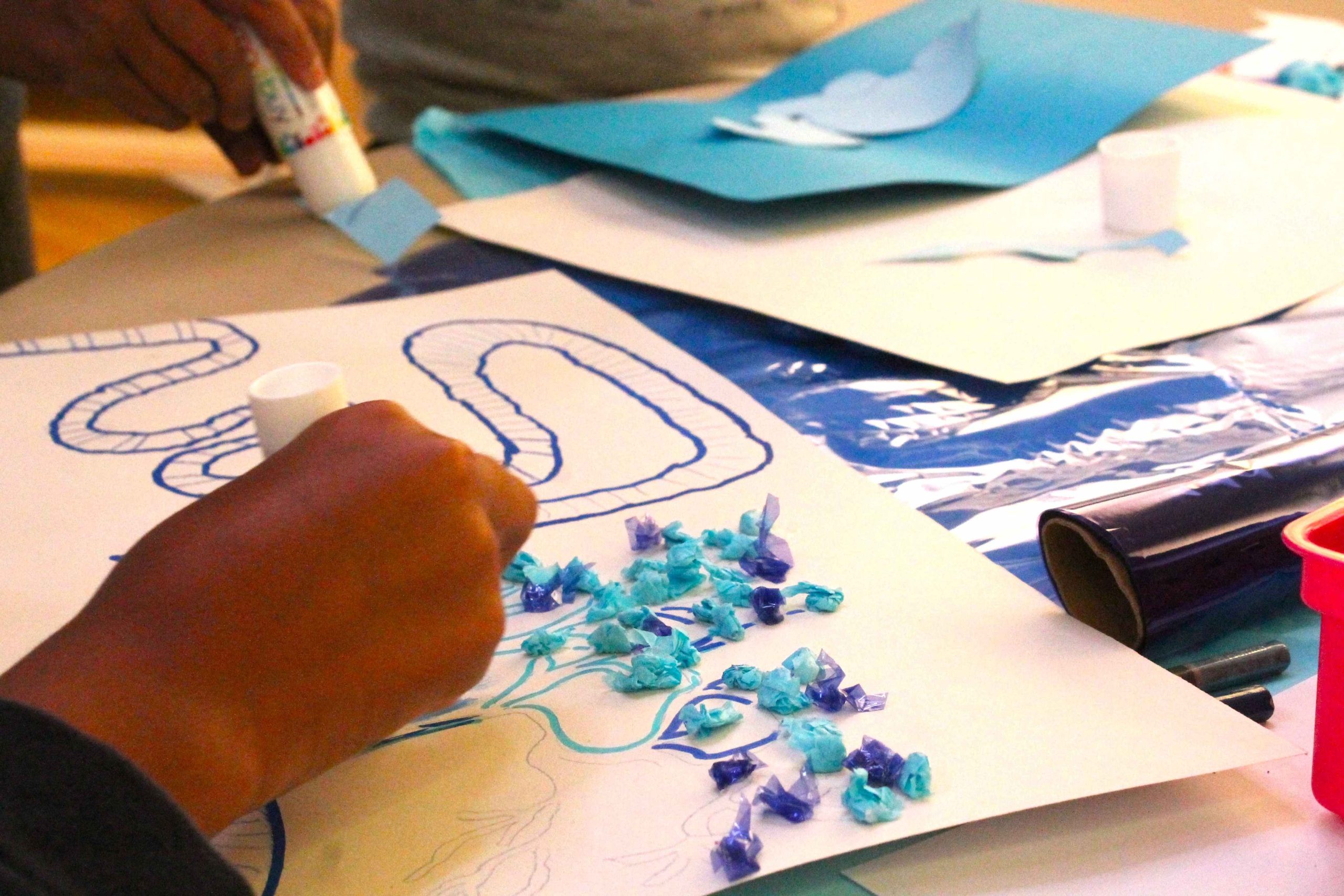A hand drawing on a white pieces of paper with blue pen to create squiggles. Glued next to it are crushed up pieces of blue and purple paper. The table is covered in paper and art materials.