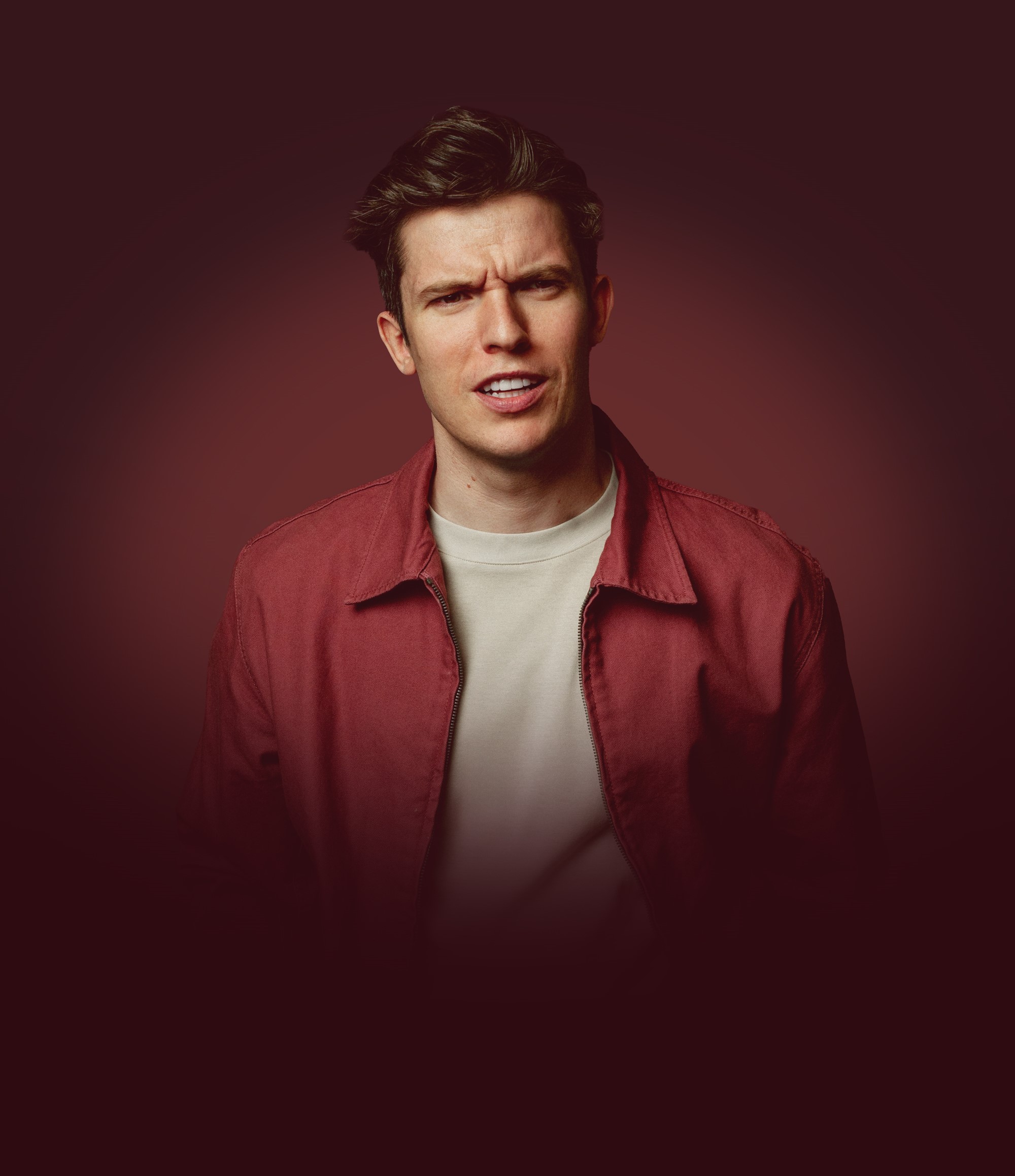 A white man looking confused in a red jacket and white t-shirt in a smoky brown background.