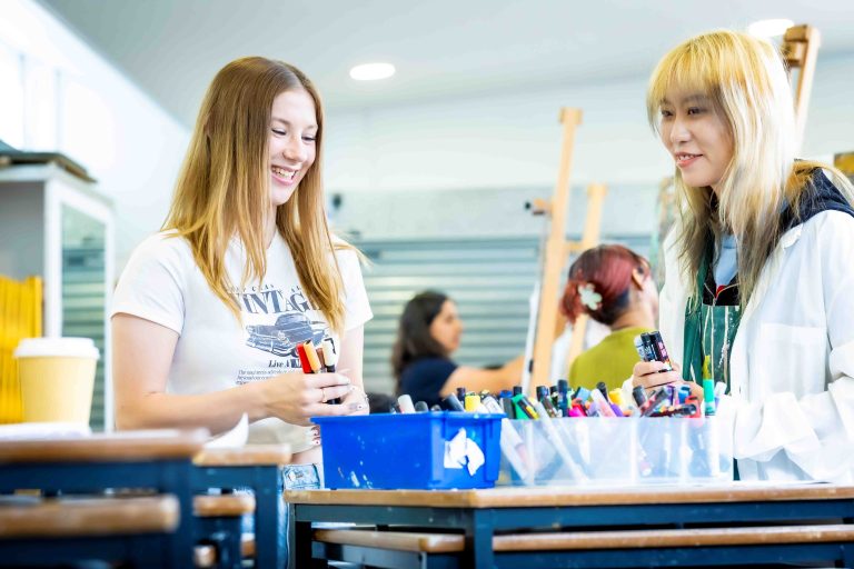 Two students smiling in a art studio, gathering art materials together.