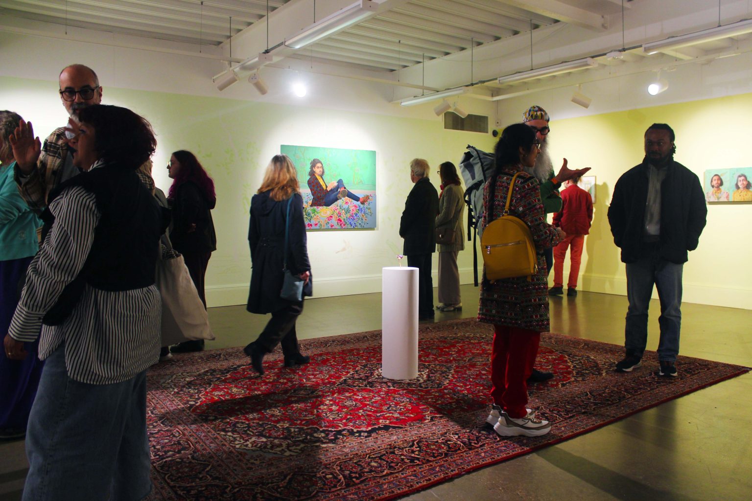 A range of people in a gallery space, with large scale oil painitngs on the wall, a large persian-style rug on the floor, and a circlular plinth in the middle of the room holding a metal sculpture of a flower.