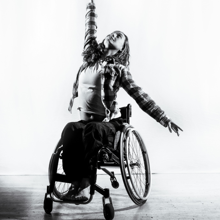 A black and white image of a woman in a wheelchair dancing with her left arm raised high.